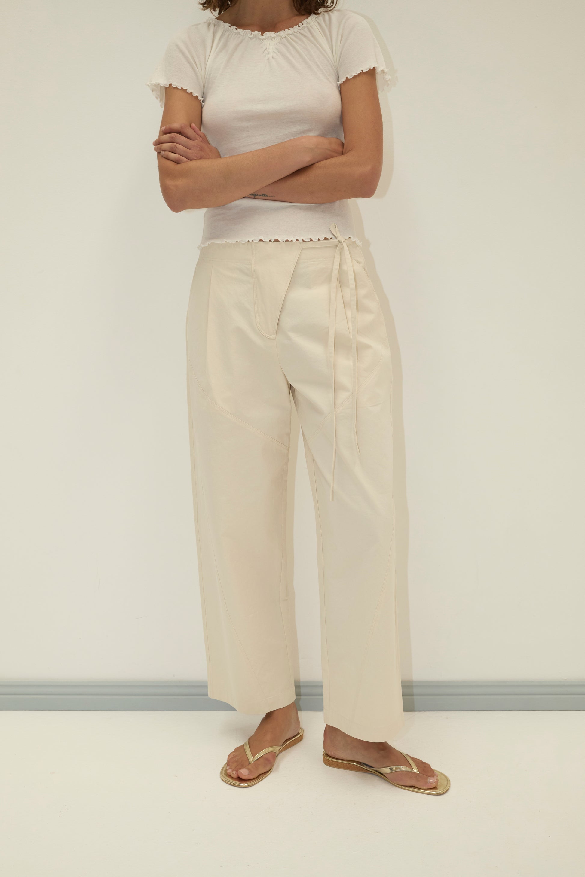 A person stands against a plain background, arms crossed, wearing the Flag Tie Pant - Off White with a white short-sleeve shirt and gold flip-flops. Their head is out of frame.