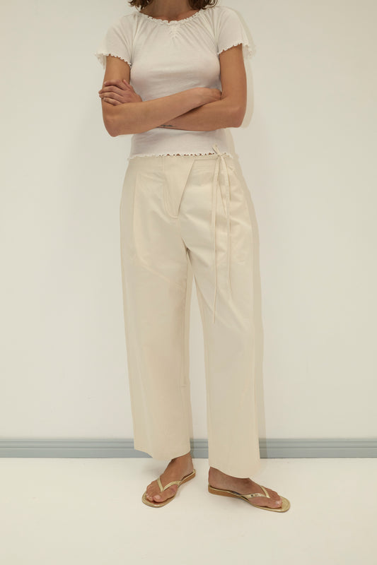 A person stands against a plain background, arms crossed, wearing the Flag Tie Pant - Off White with a white short-sleeve shirt and gold flip-flops. Their head is out of frame.