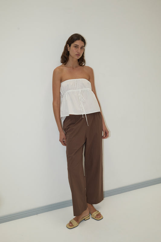 A woman with long hair and a neutral expression stands against a white wall, wearing Deiji Studios’ strapless cotton top - white in organic cotton poplin, loose brown pants, and sandals.