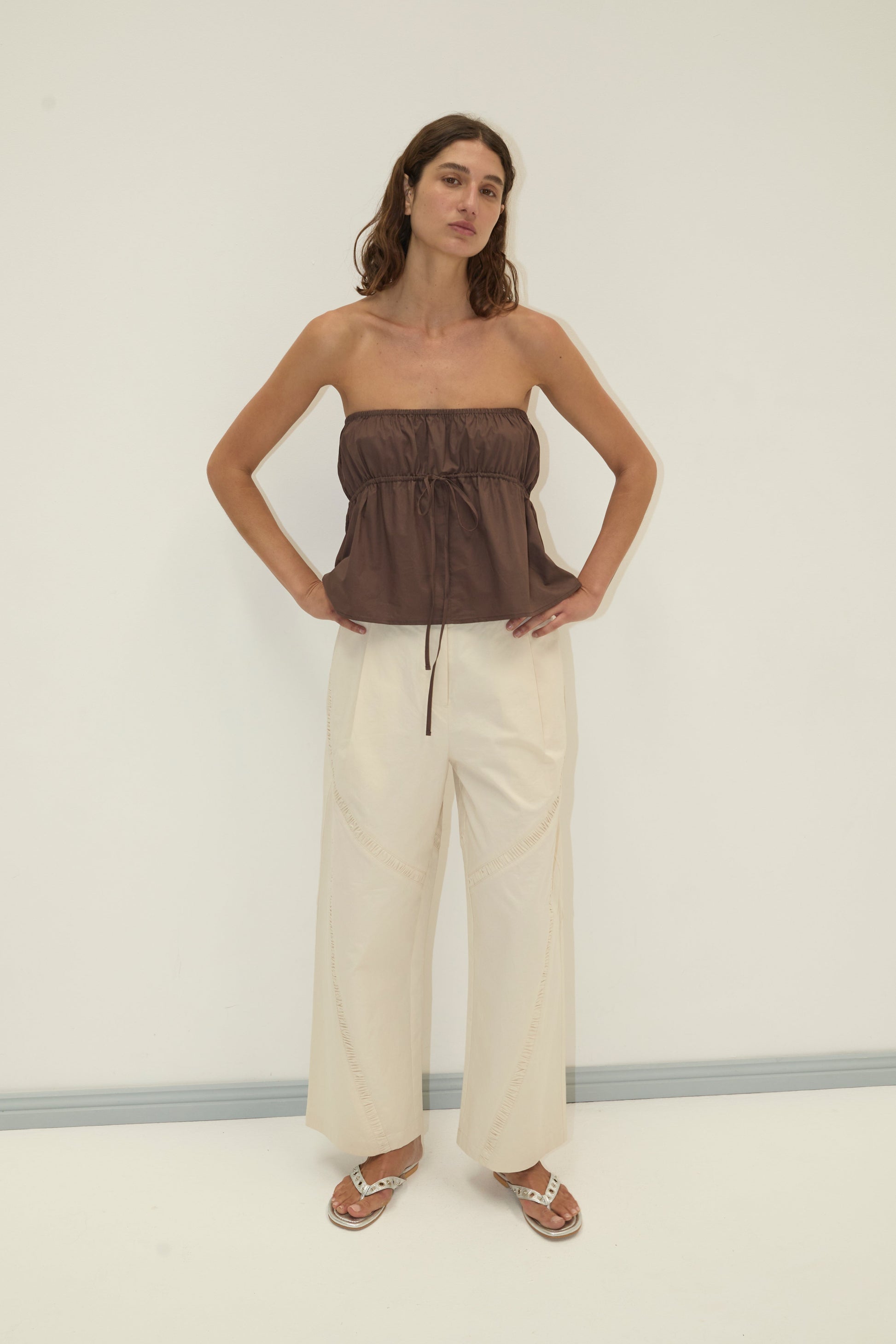 A woman poses against a plain light background in a brown Strapless Cotton Top, wide-leg cream pants, and sandals, with her hands on her hips and a neutral expression.