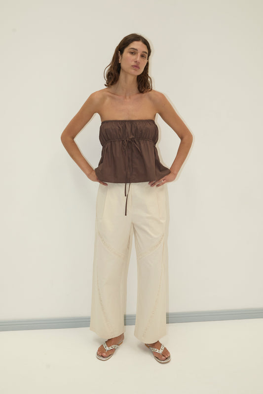 A woman poses against a plain light background in a brown Strapless Cotton Top, wide-leg cream pants, and sandals, with her hands on her hips and a neutral expression.