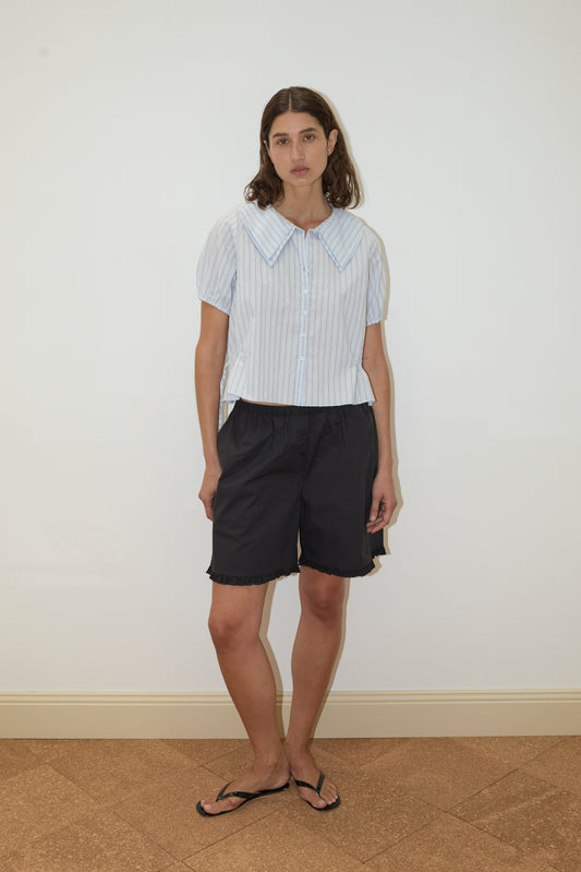 A woman with shoulder-length brown hair stands against a plain white wall, wearing a Deiji Studios Double Collar Shirt in Mineral Stripe, paired with loose black shorts and black flip-flops.