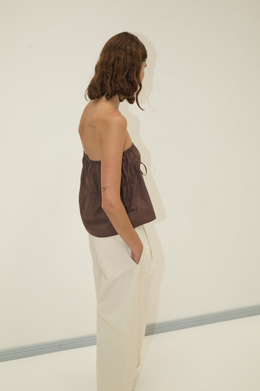 A woman with wavy brown hair stands sideways against a plain white wall, wearing the Strapless Cotton Top - Chocolate and loose, light-colored pants, her hands tucked into her pockets.