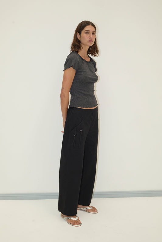 A woman with wavy brown hair stands against a plain light wall, wearing the Flounce Tee - Grey by Deiji Studios, wide-leg black pants with pockets, and sandals. She looks slightly to the side.