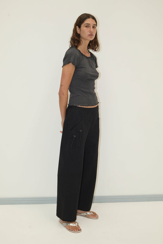 A woman with wavy brown hair stands against a plain light wall, wearing a fitted dark gray short-sleeve top and Deiji Studios Adjust Pocket Pant in black organic cotton, paired with flat sandals. She gazes slightly to the side with a neutral expression.