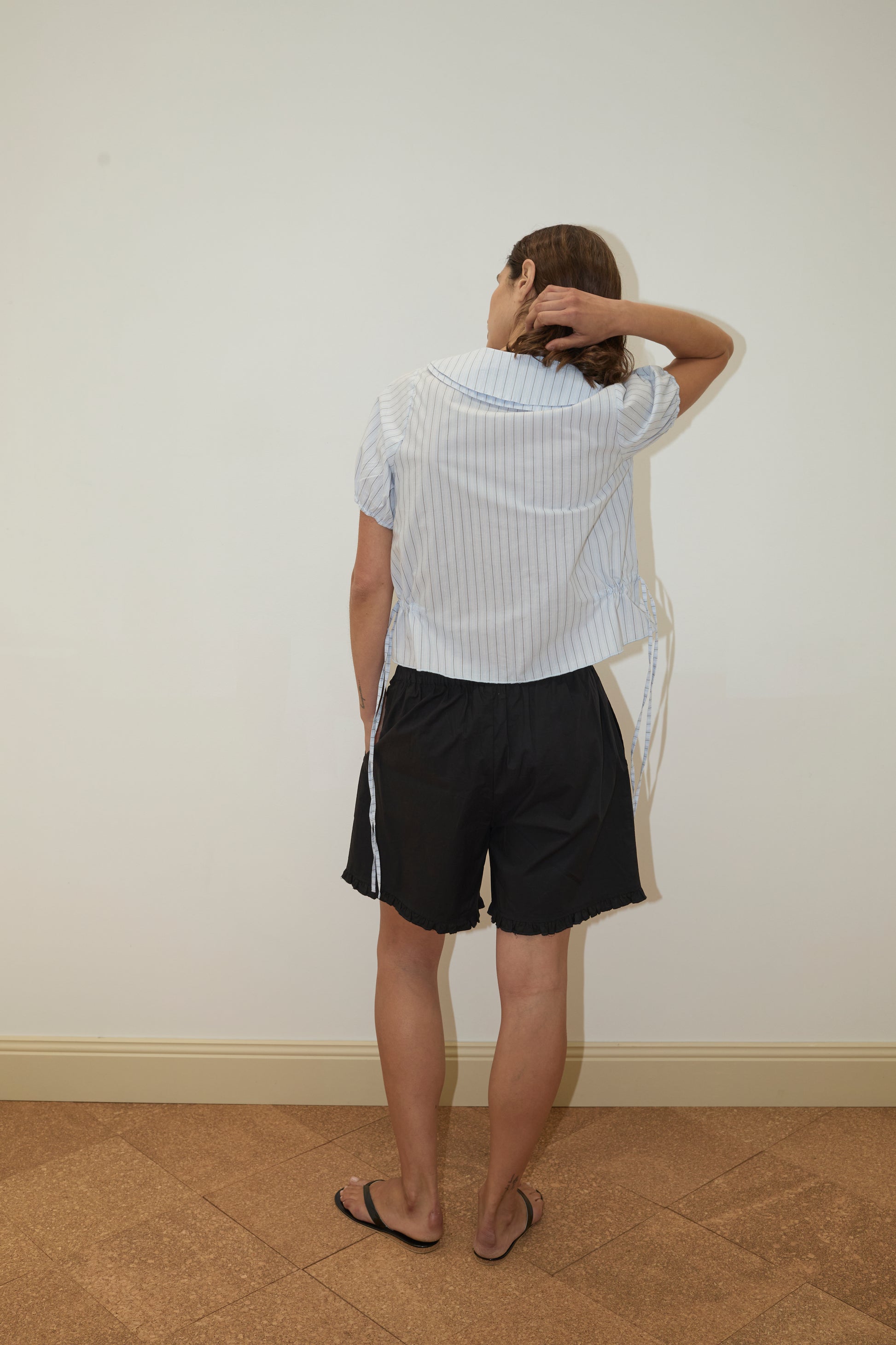 A person faces a blank wall with their back to the camera, hand on head, wearing the Deiji Studios Double Collar Shirt - Mineral Stripe, paired with black shorts and black flip-flops.