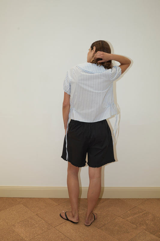 A person faces a blank wall with their back to the camera, hand on head, wearing the Deiji Studios Double Collar Shirt - Mineral Stripe, paired with black shorts and black flip-flops.