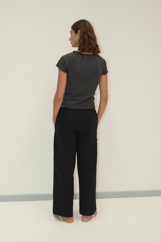 A person with shoulder-length brown hair stands with their back to the camera, wearing Deiji Studios' Adjust Pocket Pant in black and a short-sleeved organic cotton top, paired with flat shoes, against a plain light background.