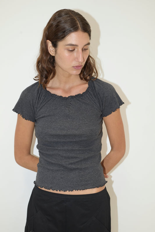 A woman with wavy brown hair wears the Flounce Tee - Grey by Deiji Studios and black pants. She stands against a plain white background with her hands behind her back, gazing down and slightly to the side.