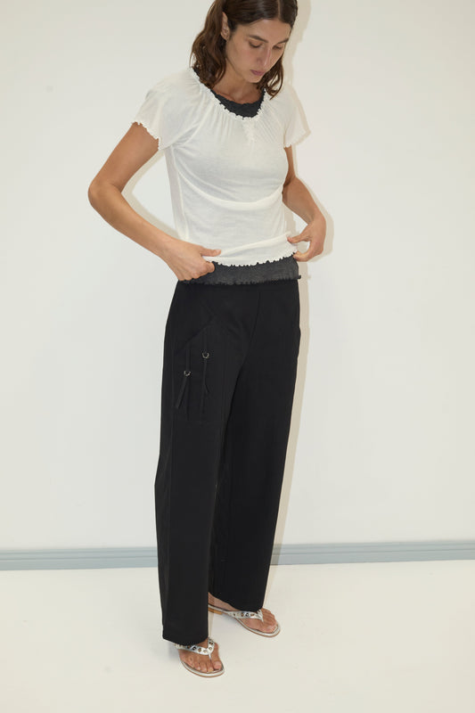 A woman stands against a white wall, wearing a Deiji Studios short-sleeve top with black trim, Adjust Pocket Pant in black, and metallic sandals. She looks down while adjusting her shirt.