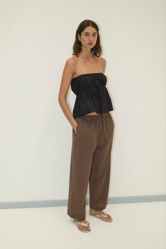A woman with wavy hair stands against a plain background, hands in her pockets, wearing a Deiji Studios strapless cotton top - black, loose brown pinstripe pants, and white sandals as she looks calmly at the camera.