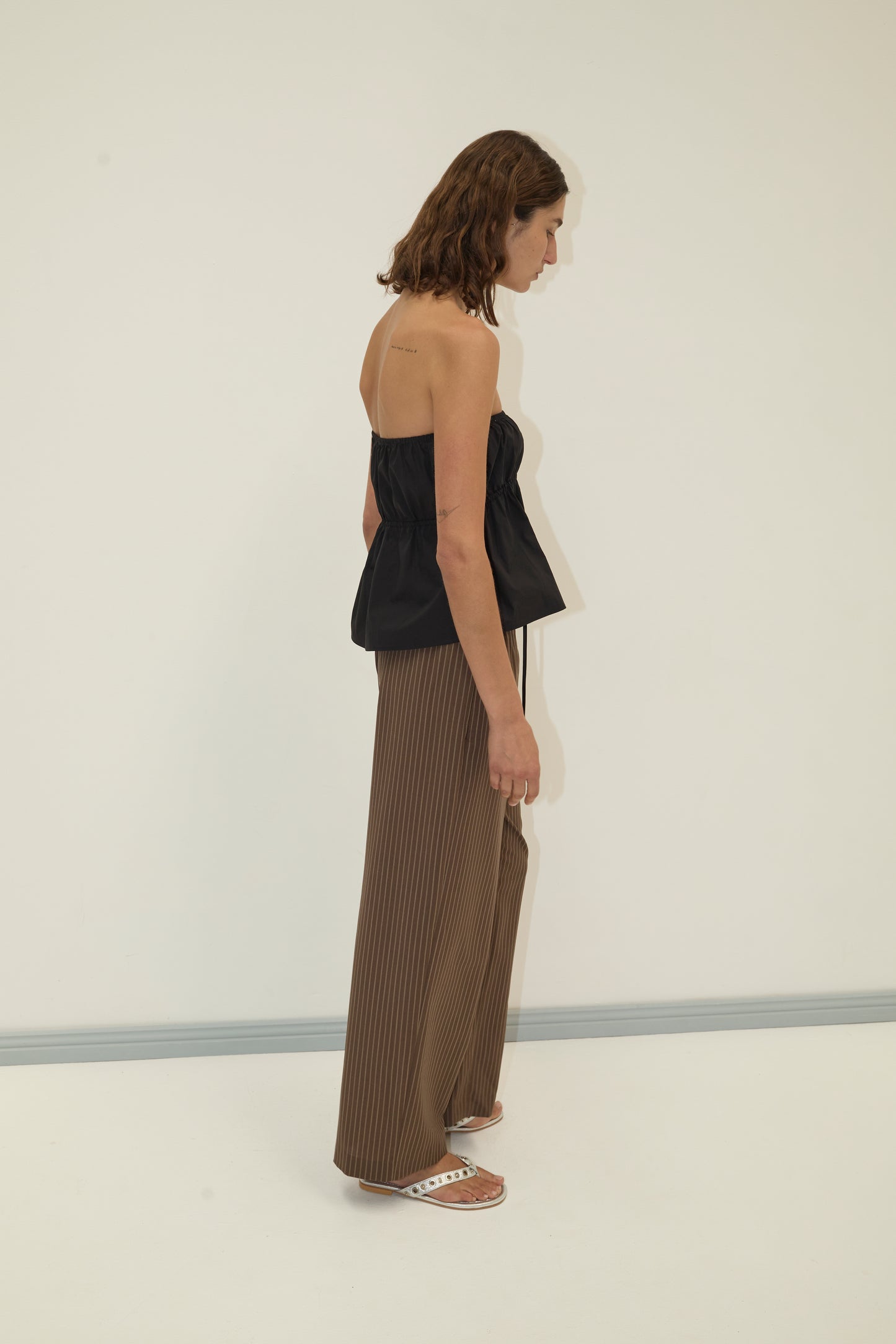 A woman with shoulder-length brown hair stands sideways against a plain white background, wearing a Deiji Studios strapless cotton top in black, brown wide-leg organic cotton poplin pants, and white slide sandals.