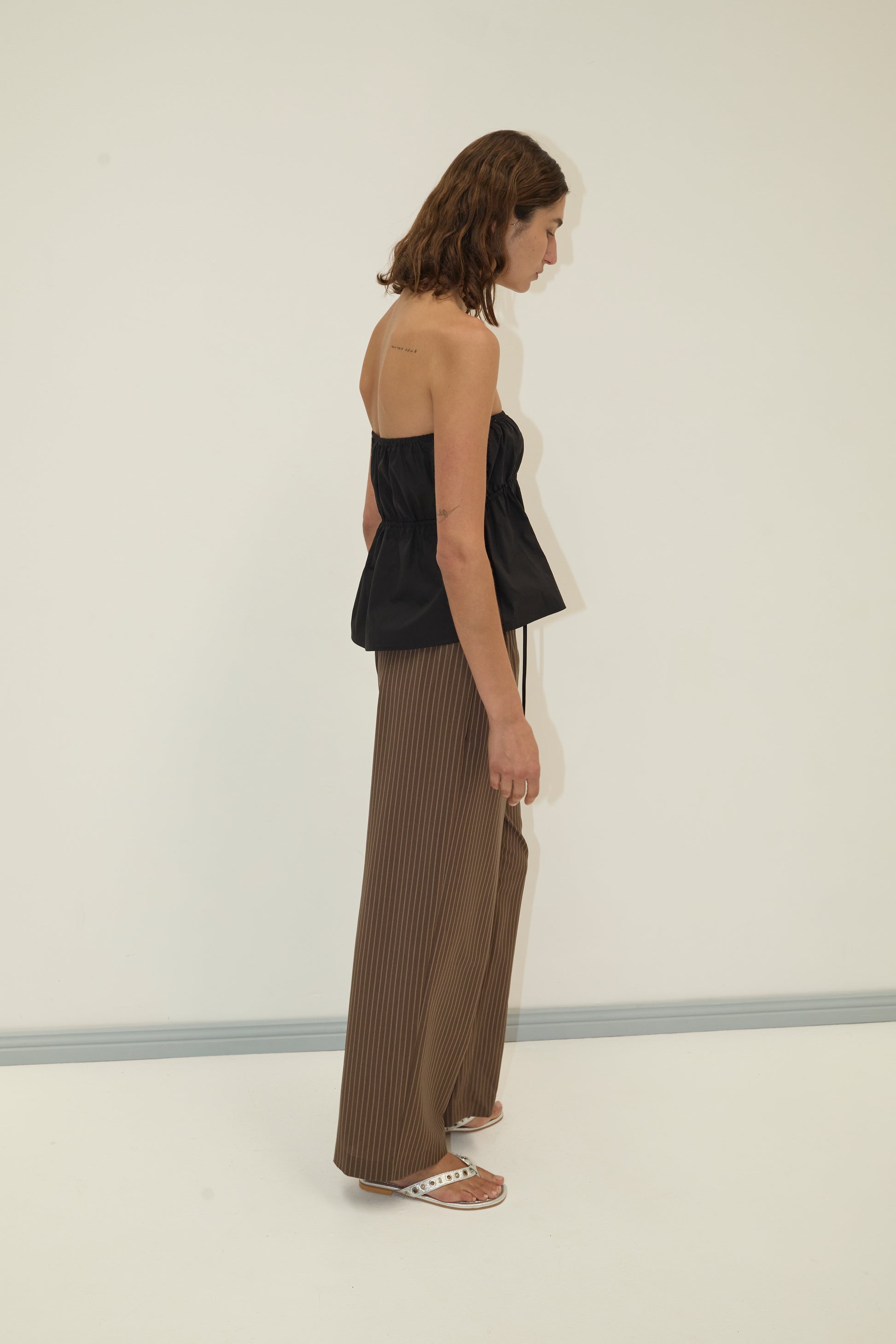 A woman with shoulder-length brown hair stands sideways against a plain white background, wearing a Deiji Studios strapless cotton top in black, brown wide-leg organic cotton poplin pants, and white slide sandals.