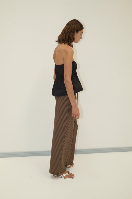 A woman with shoulder-length brown hair stands sideways against a plain white background, wearing a Deiji Studios strapless cotton top in black, brown wide-leg organic cotton poplin pants, and white slide sandals.