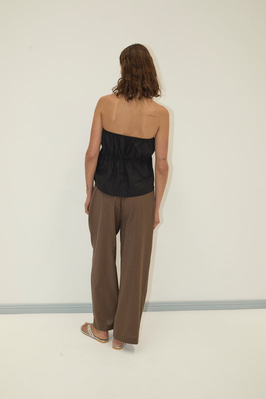 A person with shoulder-length brown hair stands facing away, wearing Deiji Studios' strapless cotton top - black, brown pinstriped wide-leg pants, and sandals against a plain light background.
