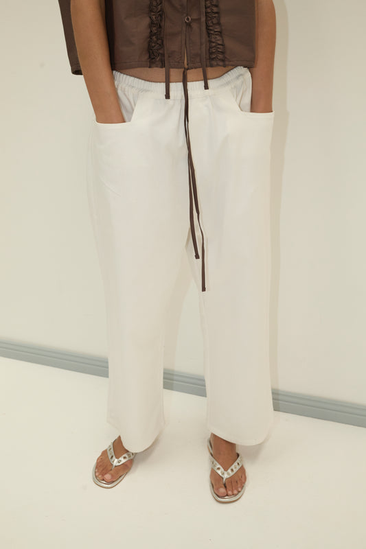 A person wears the Square Pocket Pant - White in organic cotton twill with a brown drawstring top and white sandals, standing against a light background. Their wide-leg silhouette and hands in pockets complete the relaxed look.