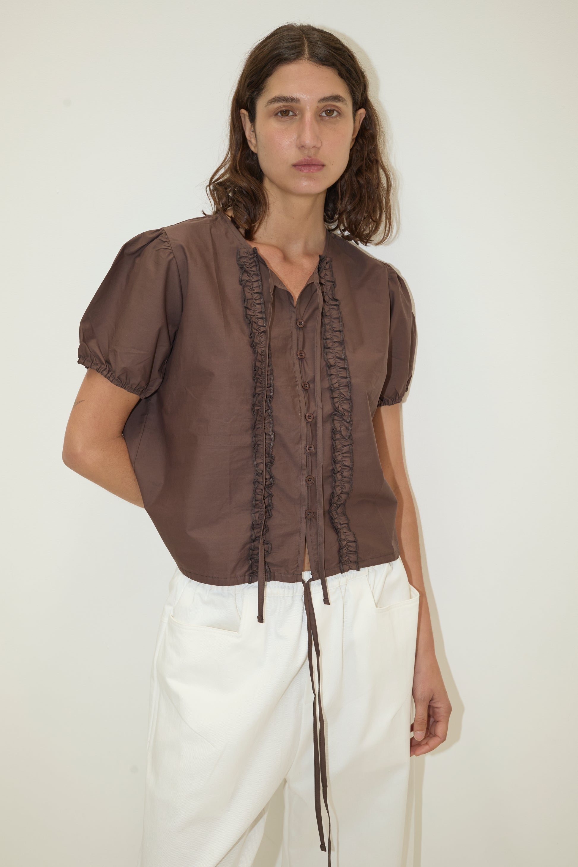 A woman with wavy brown hair wears the Double Frill Top - Chocolate by Deiji Studios and high-waisted white pants, standing against a plain light background.
