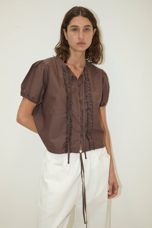 A woman with wavy brown hair wears the Double Frill Top - Chocolate by Deiji Studios and high-waisted white pants, standing against a plain light background.