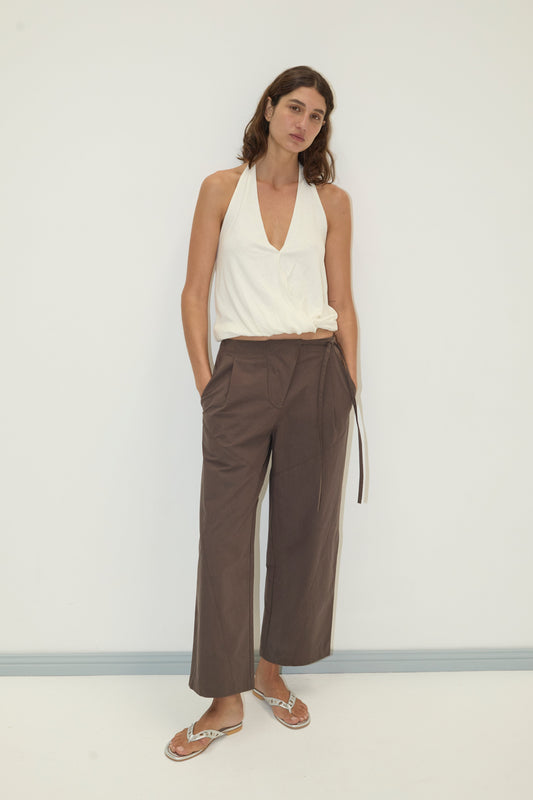 A woman with medium-length brown hair stands against a white wall, hands in her pockets, wearing the Flag Tie Pant—loose brown trousers with a belt—along with a sleeveless white halter top and white sandals.