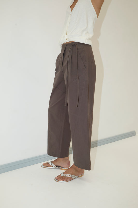 A person wears the Flag Tie Pant in Dark Brown with a sleeveless white top and metallic flip-flops, standing against a plain white wall. Their face is not visible.