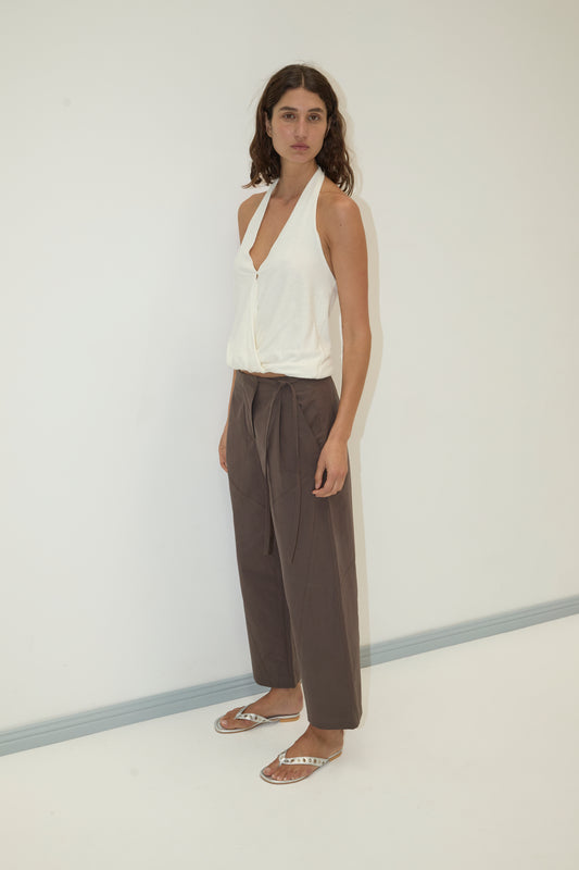 A woman stands against a plain white wall, wearing a sleeveless white top, silver sandals, and the Flag Tie Pant in Dark Brown. Her wavy brown hair and neutral expression complete the effortless look.