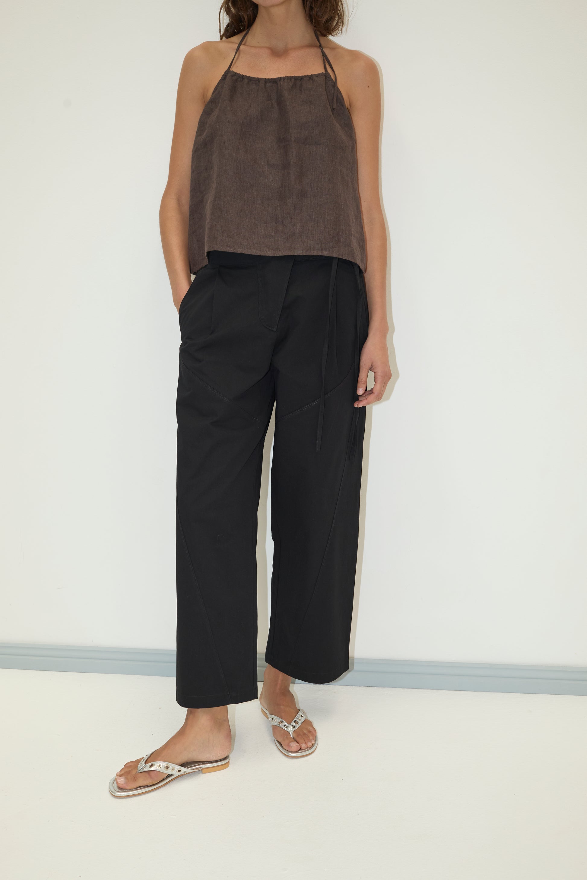 A person stands against a plain white wall, wearing loose black Flag Tie Pants with pockets, a brown halter-neck top, and tan flip-flops. Their left hand is in their pocket, and their head is not visible in the image.