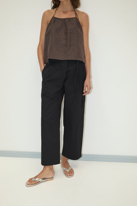 A person stands against a plain white wall, wearing loose black Flag Tie Pants with pockets, a brown halter-neck top, and tan flip-flops. Their left hand is in their pocket, and their head is not visible in the image.