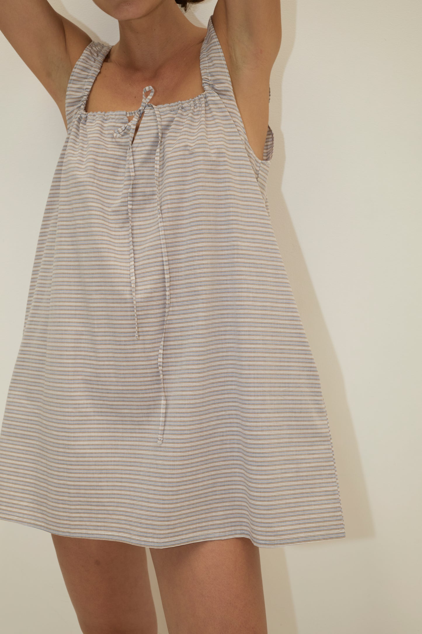 A person wearing The Bow Dress - Horizontal Stripe, a sleeveless, loose-fit mini dress in organic cotton with thin stripes, stands against an off-white background with arms raised and face partially out of frame.