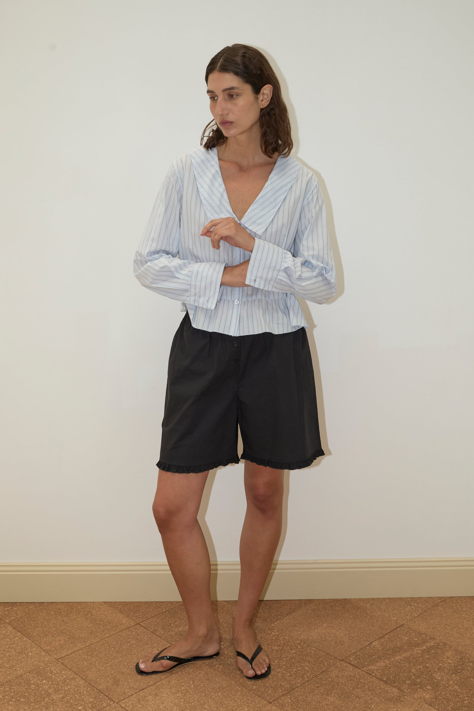 A person stands indoors against a plain white wall, wearing the Sweep Shirt - Mineral Stripe by Deiji Studios, black shorts, and black flip-flops. They have short brown hair and are looking to the side with arms loosely crossed.