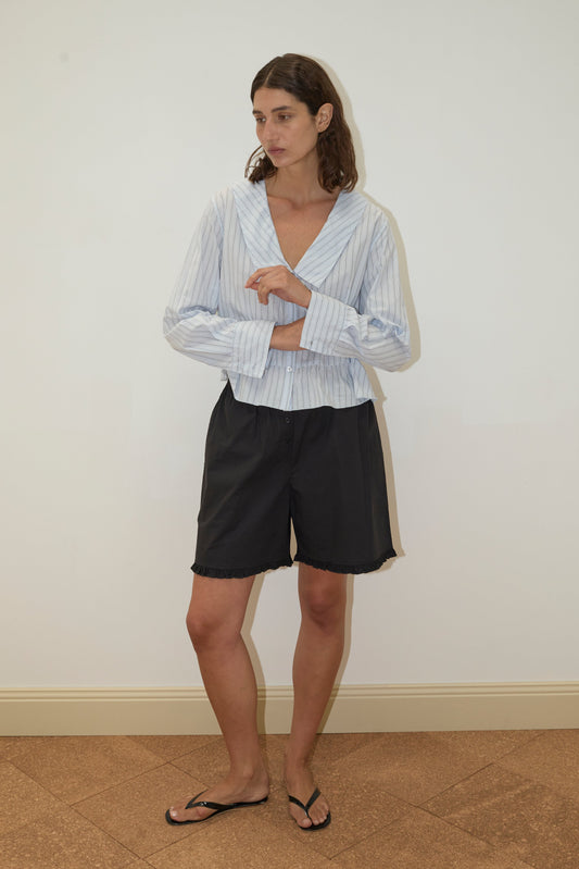 A person stands indoors against a plain white wall, wearing the Sweep Shirt - Mineral Stripe by Deiji Studios, black shorts, and black flip-flops. They have short brown hair and are looking to the side with arms loosely crossed.