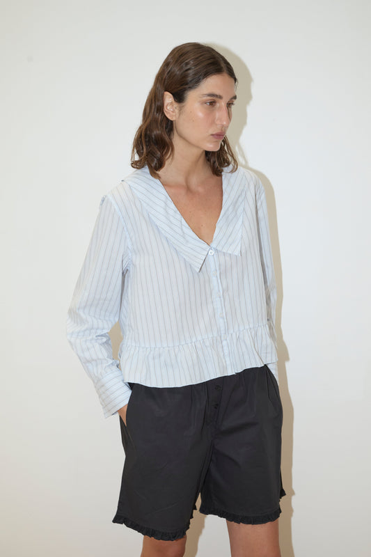 A woman with wavy brown hair stands against a plain white background, wearing the Deiji Studios Sweep Shirt - Mineral Stripe with light blue and white stripes and black shorts. She has one hand in her pocket and gazes slightly to the side.