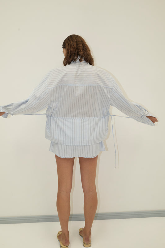 A person with wavy hair stands barefoot on a light floor, arms outstretched, wearing Deiji Studios' Room Shirt - Mineral Stripe and matching shorts, facing a white wall in a softly lit room.