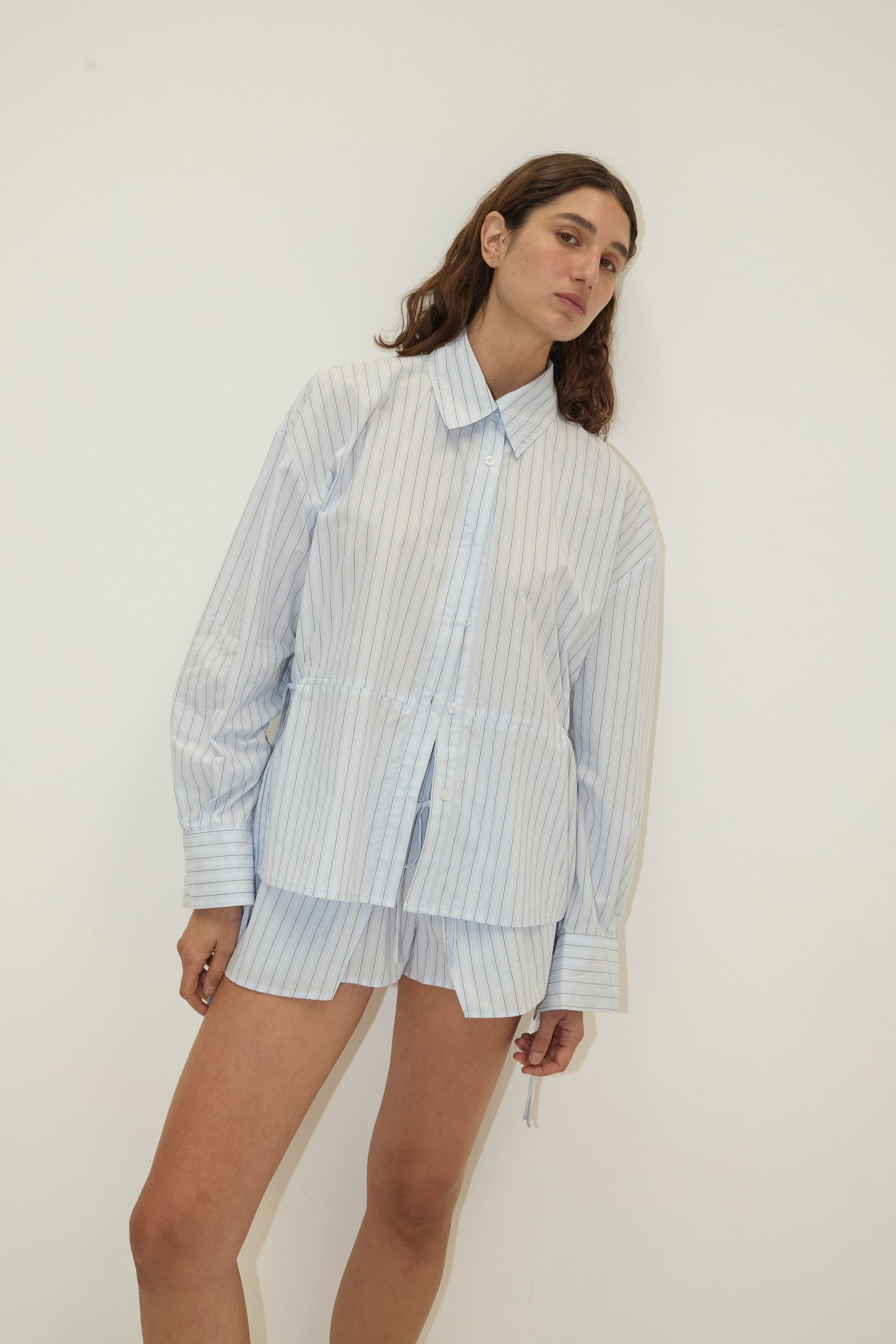 A woman with a calm expression and tilted head stands against a white background wearing the Deiji Studios Room Shirt in Mineral Stripe, paired with matching shorts.