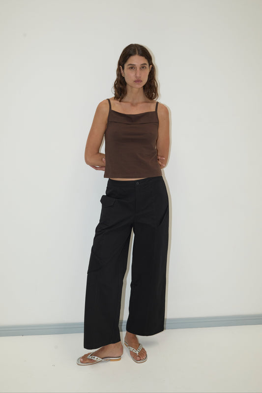 A woman with long brown hair stands against a white wall, wearing the Pocket Pant - Black with a brown sleeveless top and flip-flops. She appears relaxed, arms at her sides, and has a neutral expression.