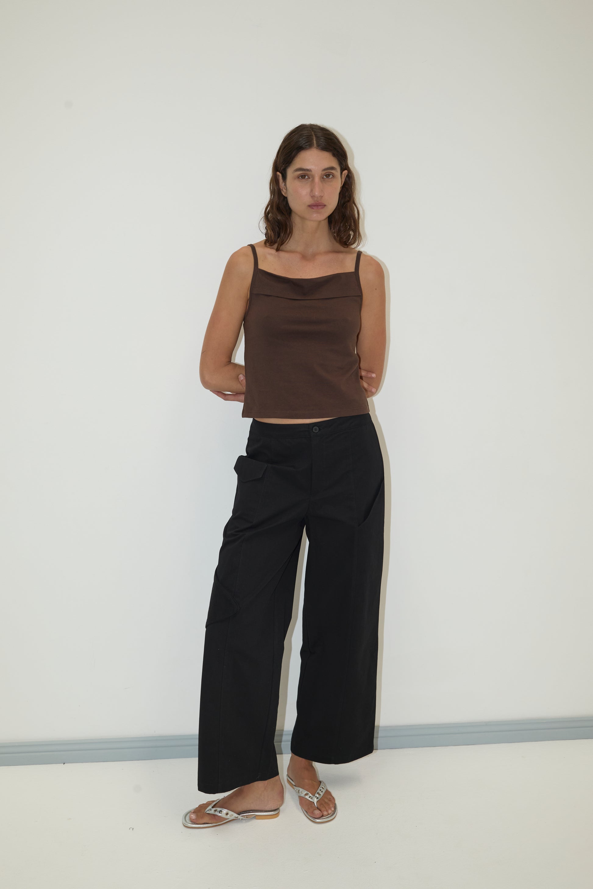 A woman with wavy brown hair stands against a white wall, wearing the Simple Tank - Caribou by Deiji Studios, loose black pants, and flip-flops. Her arms are relaxed behind her back and she has a neutral expression.