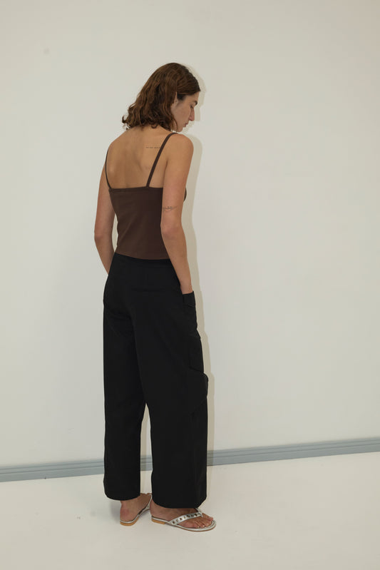 A woman with short brown hair stands facing away, wearing the Pocket Pant - Black and a dark brown spaghetti strap top with sandals, against a plain light-colored wall.