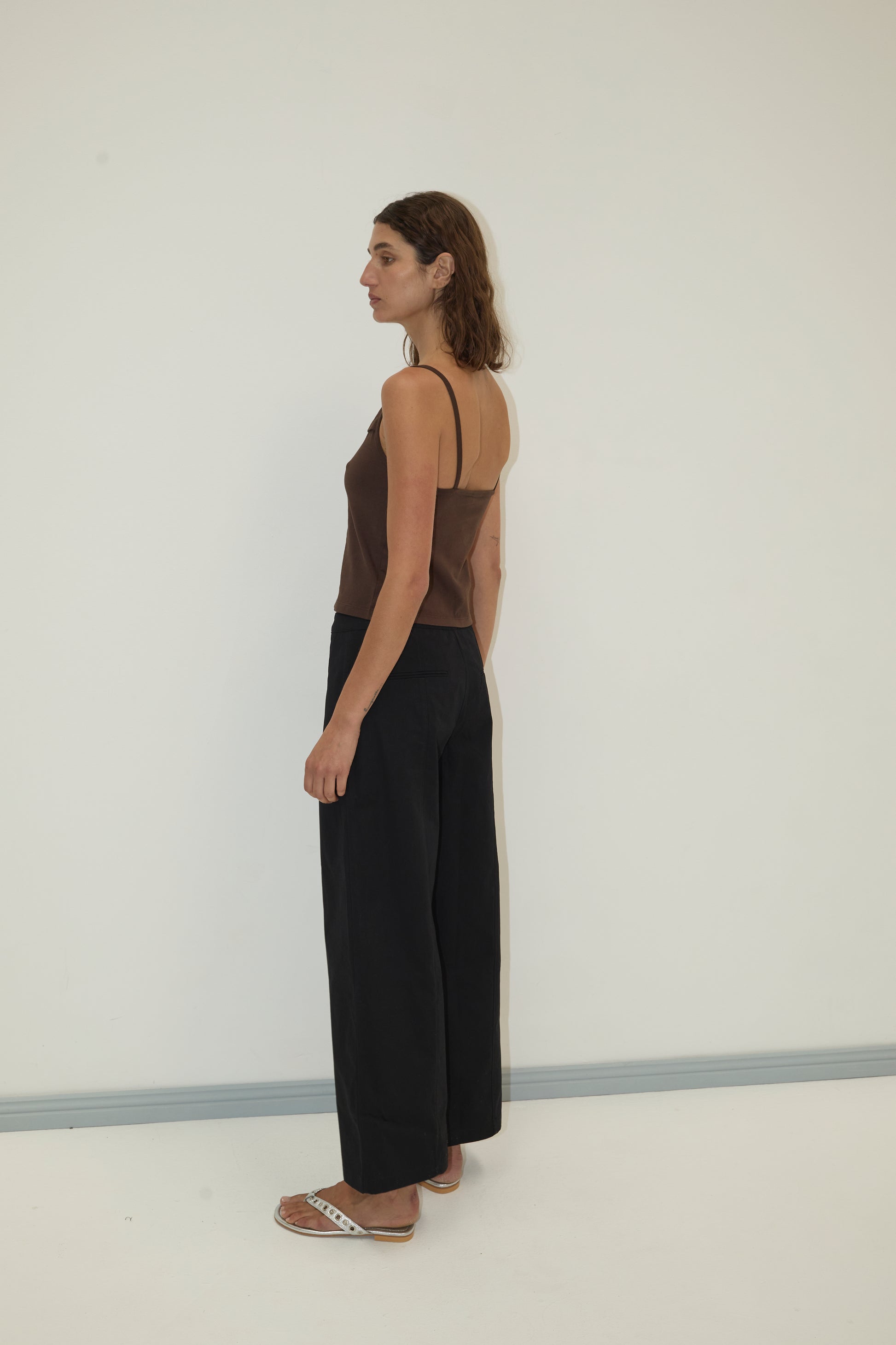 A woman with shoulder-length brown hair stands sideways against a plain white wall, wearing the Simple Tank - Caribou from Deiji Studios, loose black pants, and white sandals, her posture relaxed.