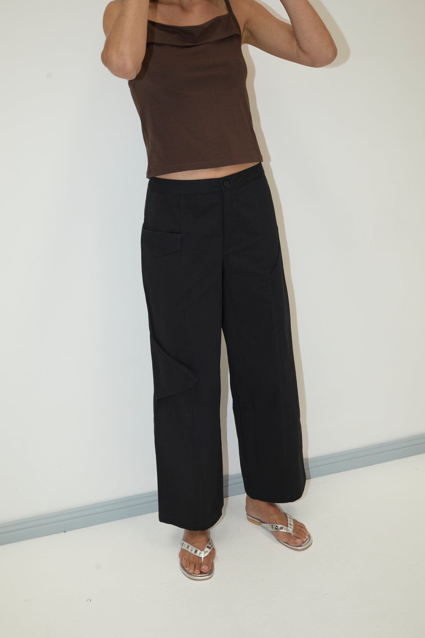 Someone stands against a plain white wall wearing a brown sleeveless top, Pocket Pant - Black with cargo pocket details, and silver sandals. Their head is cropped out of the frame.