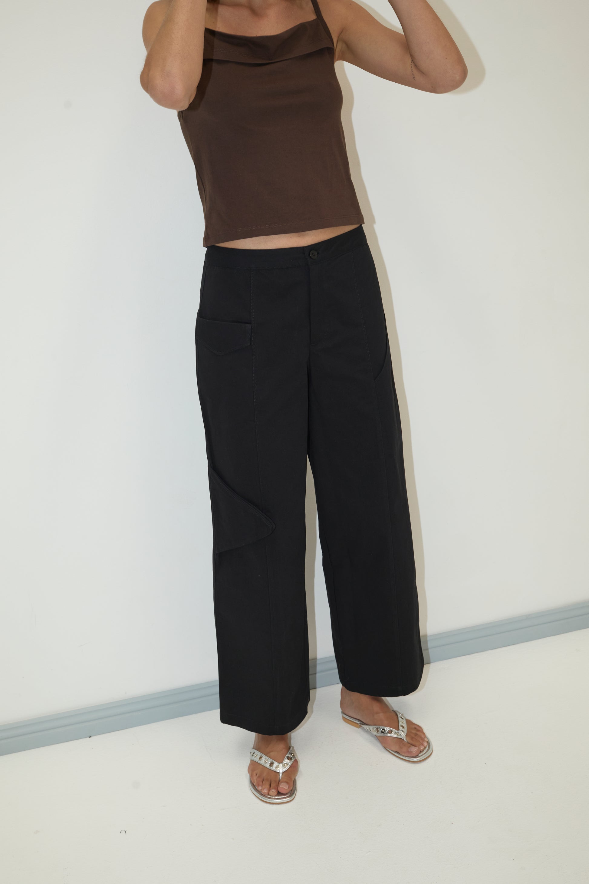 Someone stands against a plain white wall wearing a brown sleeveless top, Pocket Pant - Black with cargo pocket details, and silver sandals. Their head is cropped out of the frame.