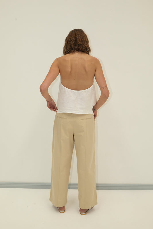 A person with wavy hair stands facing away, wearing a white backless top and Deiji Studios Pocket Pant - Stone, made from organic cotton twill, in beige against a plain light background.