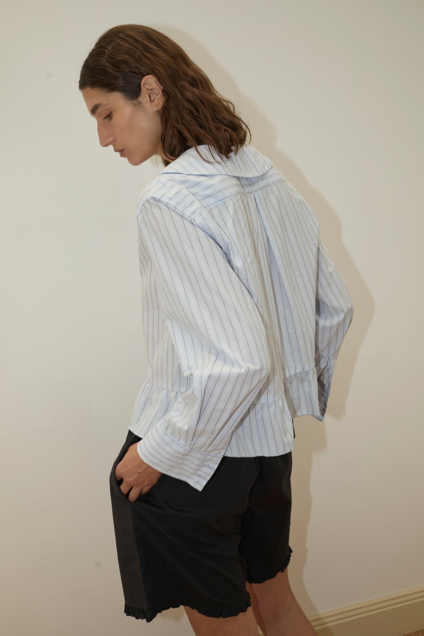 A person with shoulder-length brown hair stands sideways against a plain light wall, wearing a loose, light blue pinstriped shirt and the Flounce Short - Black from Deiji Studios.