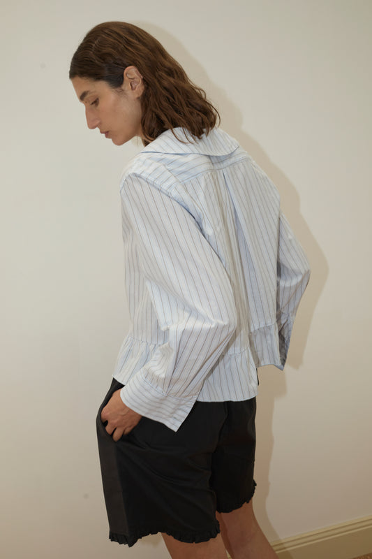 A person with shoulder-length brown hair stands sideways against a plain light wall, wearing a loose, light blue pinstriped shirt and the Flounce Short - Black from Deiji Studios.