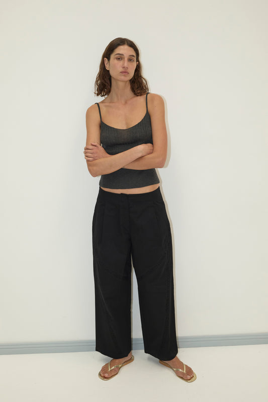 A woman with wavy brown hair stands against a white wall, arms crossed. She wears a black tank top, gold sandals, and the Channel Pant in black, looking calmly at the camera.