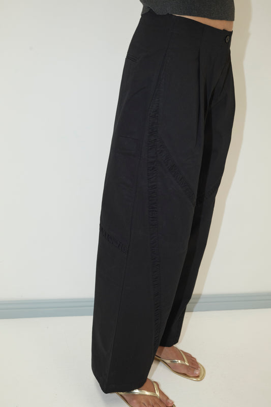 Someone stands in side profile against a plain light wall, wearing the Channel Pant - Black. The photo, cropped from chest to feet, highlights the relaxed straight-leg fit paired with gold sandals.