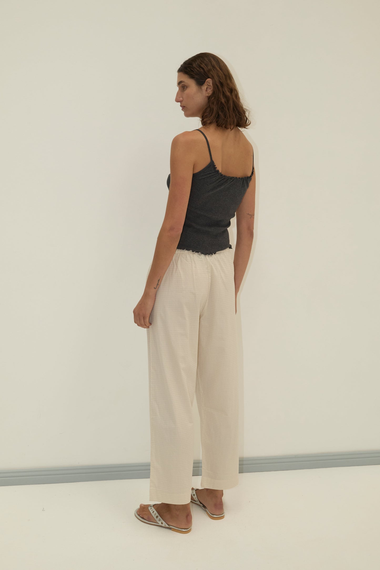 A person with shoulder-length brown hair stands in a neutral pose, facing sideways, wearing a dark gray camisole, Deiji Studios Ease Trouser - Bone Check, and sandals against a plain light-colored background.