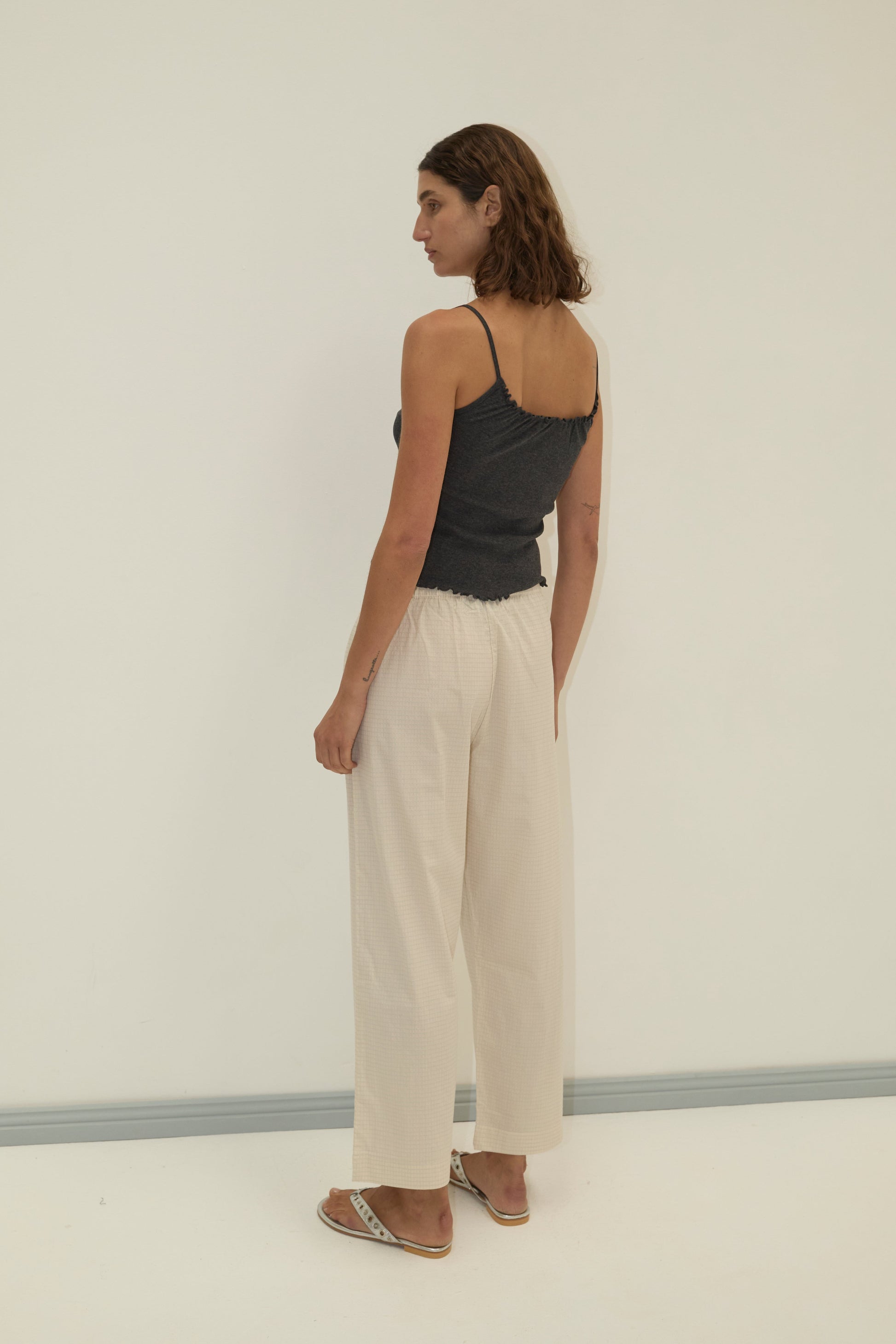 A person with shoulder-length brown hair stands in a neutral pose, facing sideways, wearing a dark gray camisole, Deiji Studios Ease Trouser - Bone Check, and sandals against a plain light-colored background.
