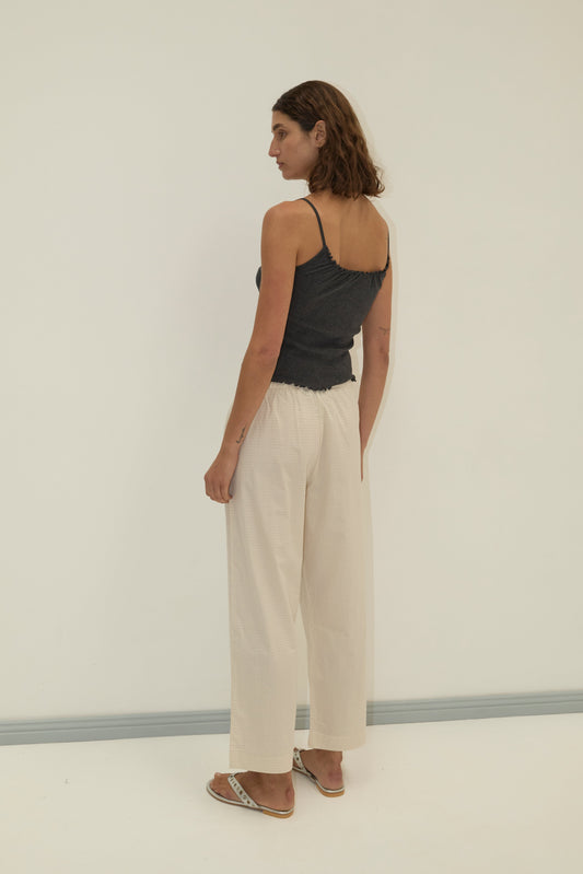 A person with shoulder-length brown hair stands in a neutral pose, facing sideways, wearing a dark gray camisole, Deiji Studios Ease Trouser - Bone Check, and sandals against a plain light-colored background.