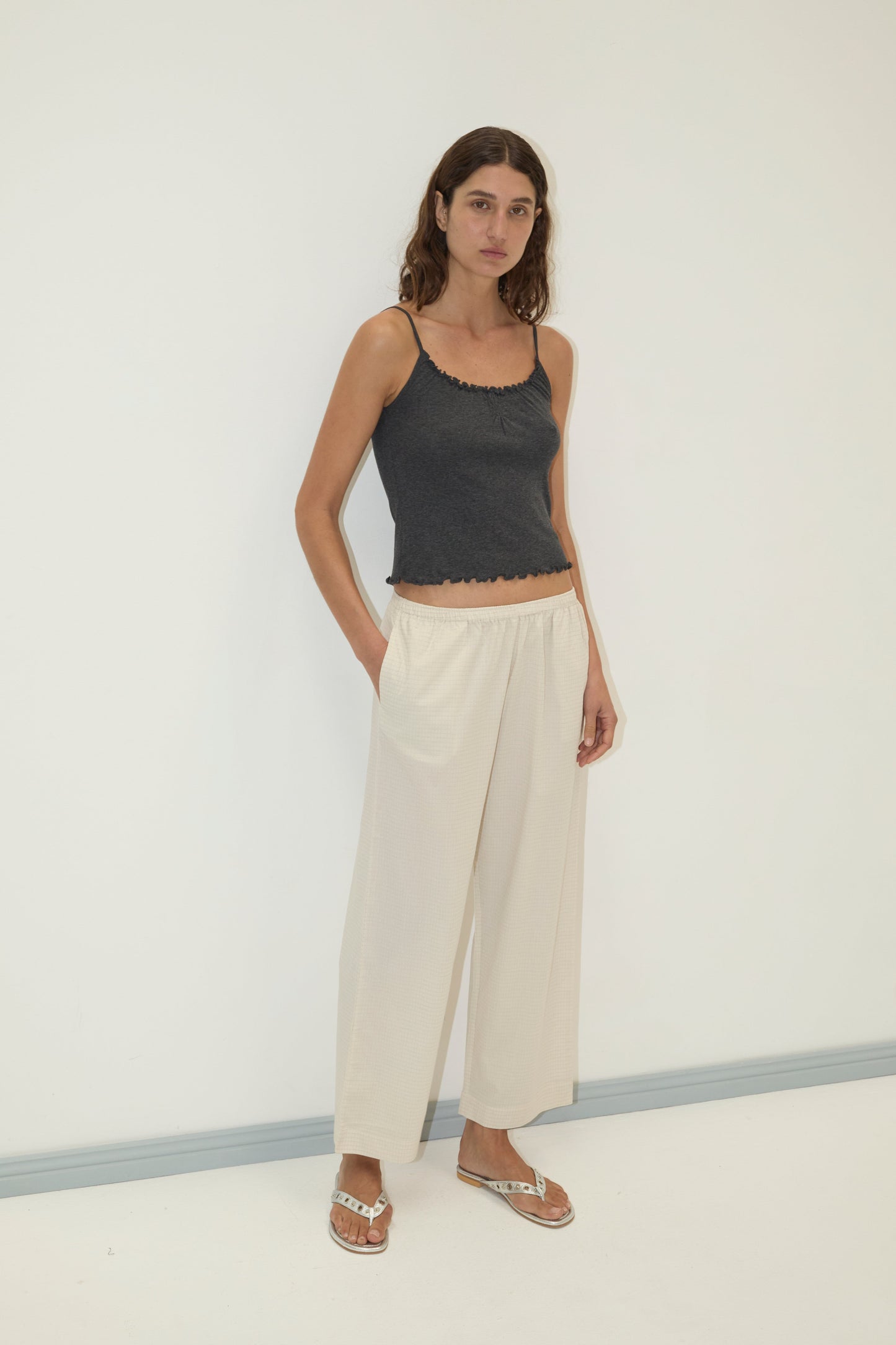 A woman with long brown hair stands against a white wall, wearing the Ease Trouser in cream, a dark gray spaghetti strap top, and sandals. With a neutral expression and one hand in her pocket, she exudes effortless style.
