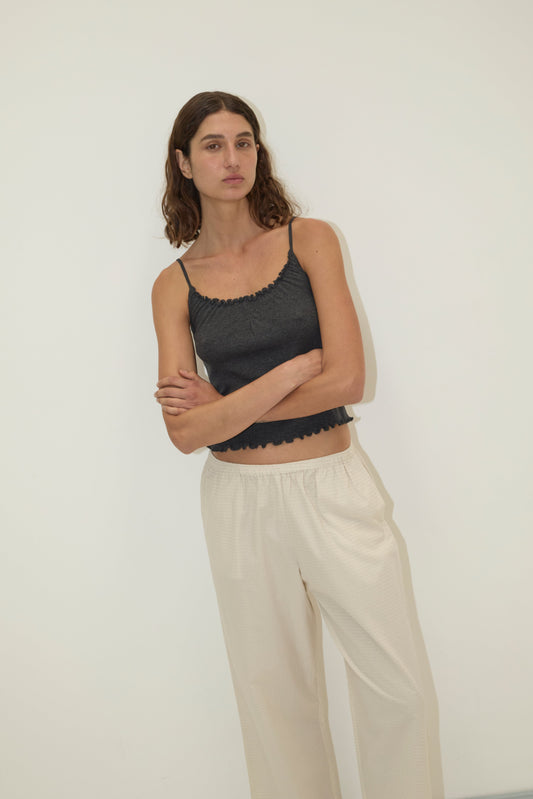 A woman with wavy brown hair stands against a white background, wearing the Flounce Tank - Grey and loose, light pants. Her arms are crossed, and she has a neutral expression.