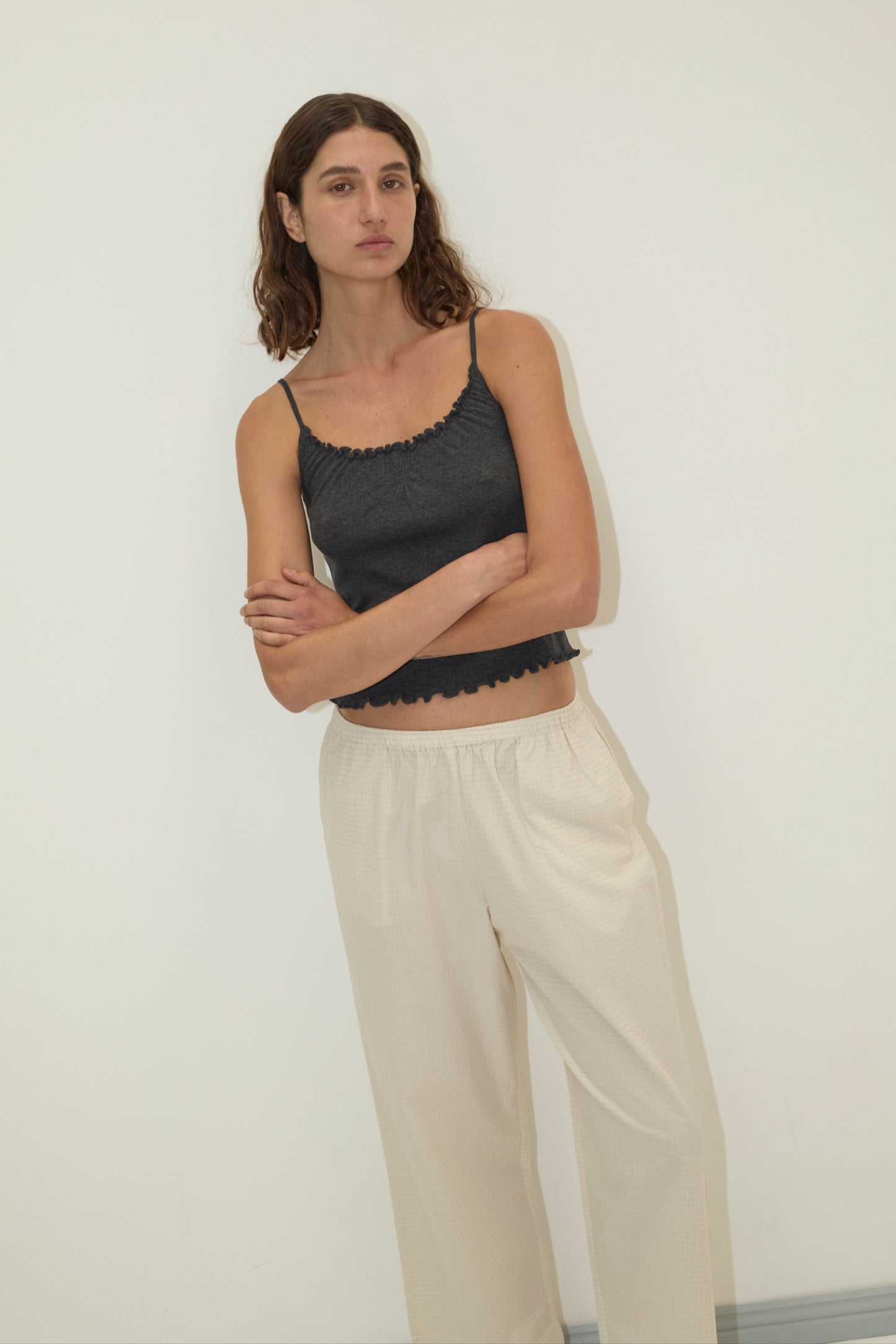 A woman with wavy brown hair stands against a plain white wall, wearing a dark gray sleeveless crop top and Deiji Studios' Ease Trouser in Bone Check, her arms loosely crossed and her expression neutral.